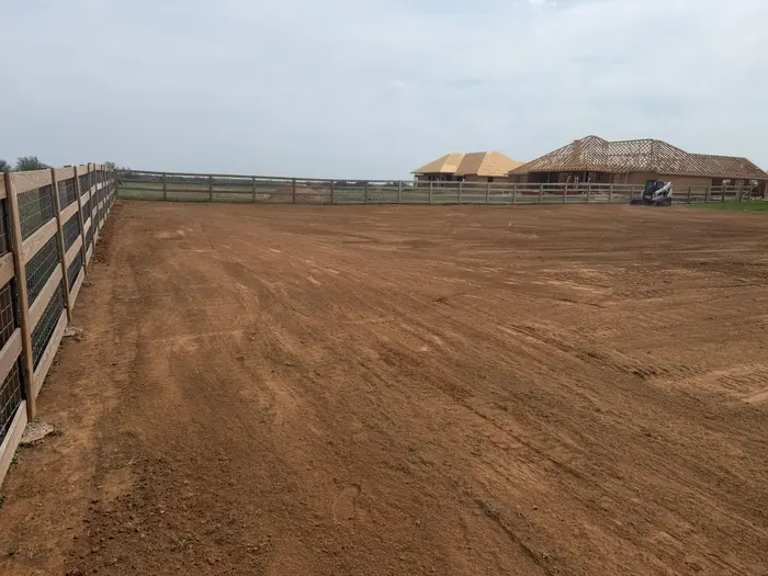 Freshly leveled arena pad inside a new board fence, home under construction behind