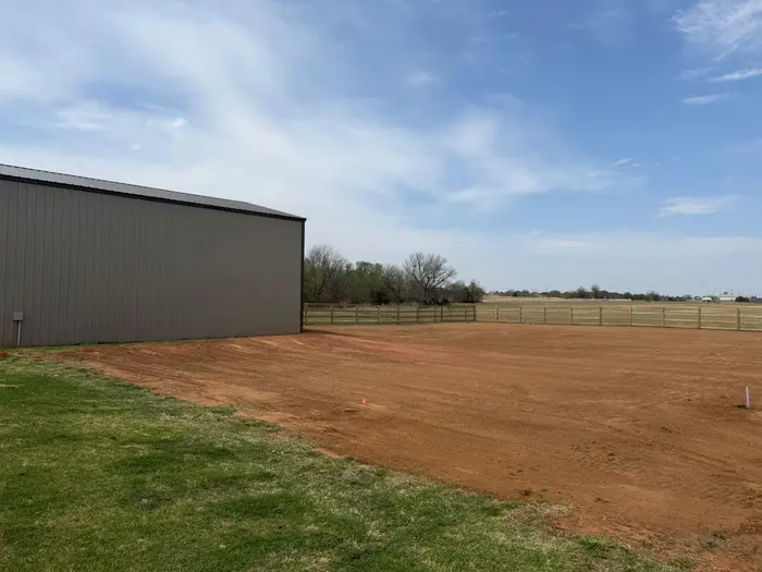 Leveled red dirt pad beside a gray metal barn with fenced pasture behind