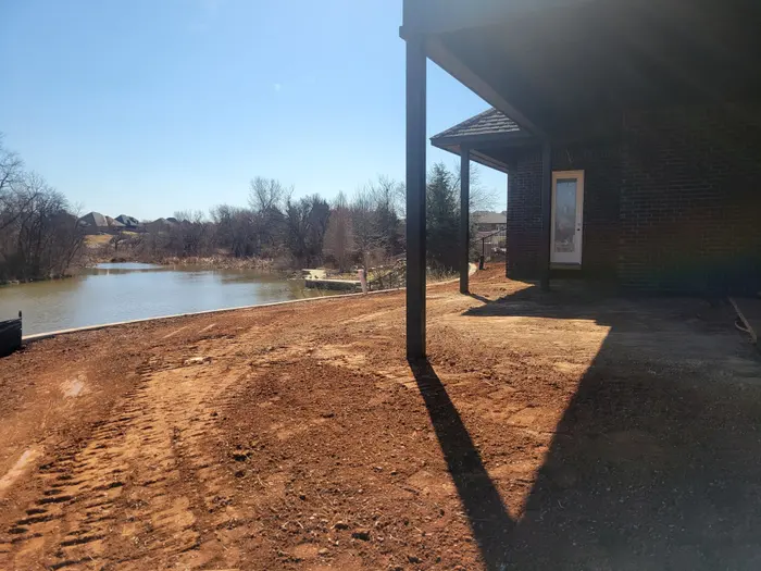 Red dirt grade worked around a lakeside home's covered porch, pond behind